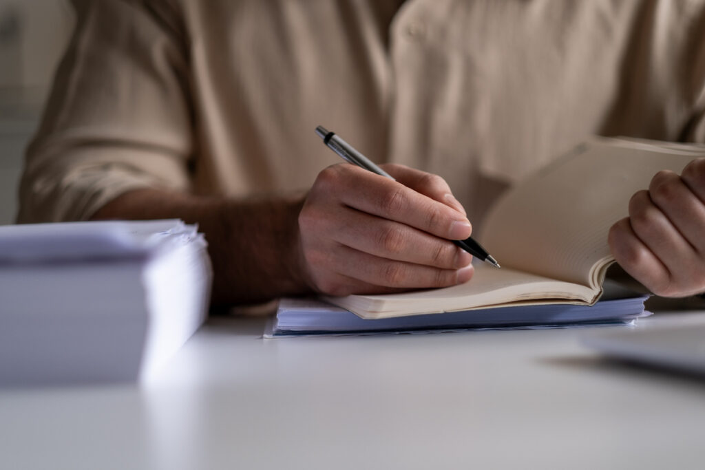 A close-up image of a man writing in a notebook with a pen 